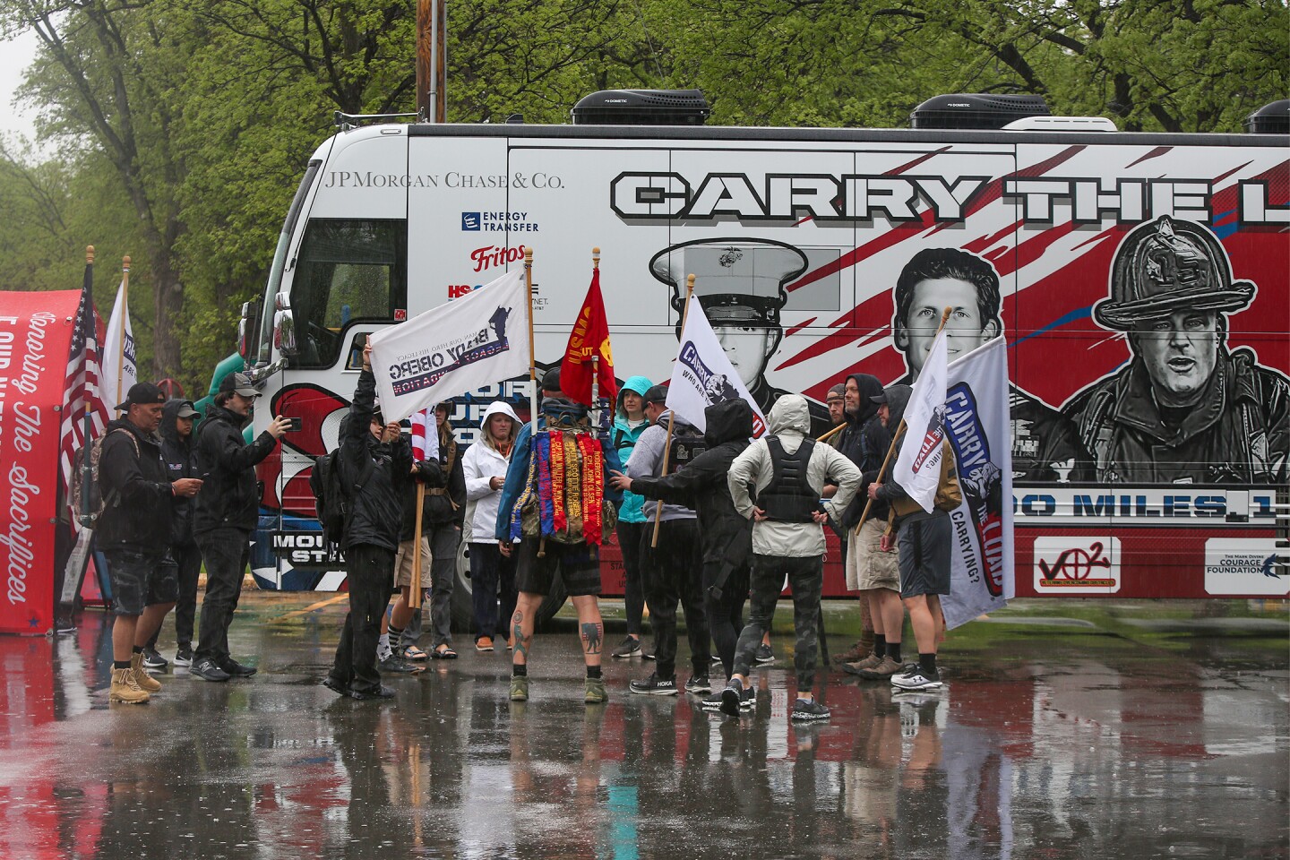 A group of people carrying flags stands in a group in a parking lot on a rainy day. They stand in a large bus which is red and features depictions of military and first responders.